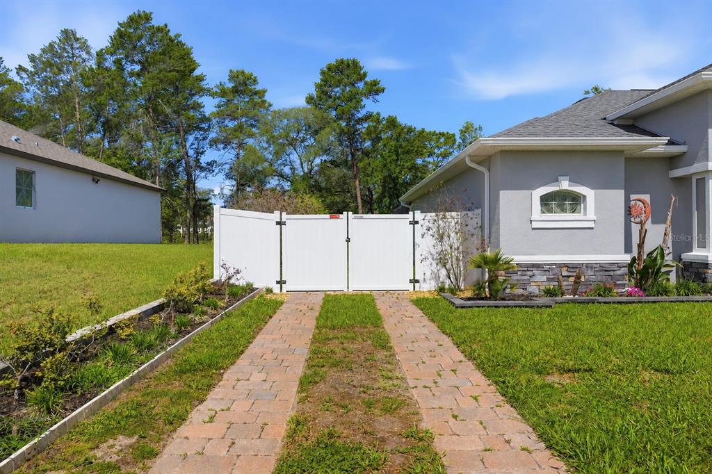 A paved driveway leads to a white vinyl gate beside a house with stone accents — 13079 HARRIS HAWK ROAD, BROOKSVILLE, FL