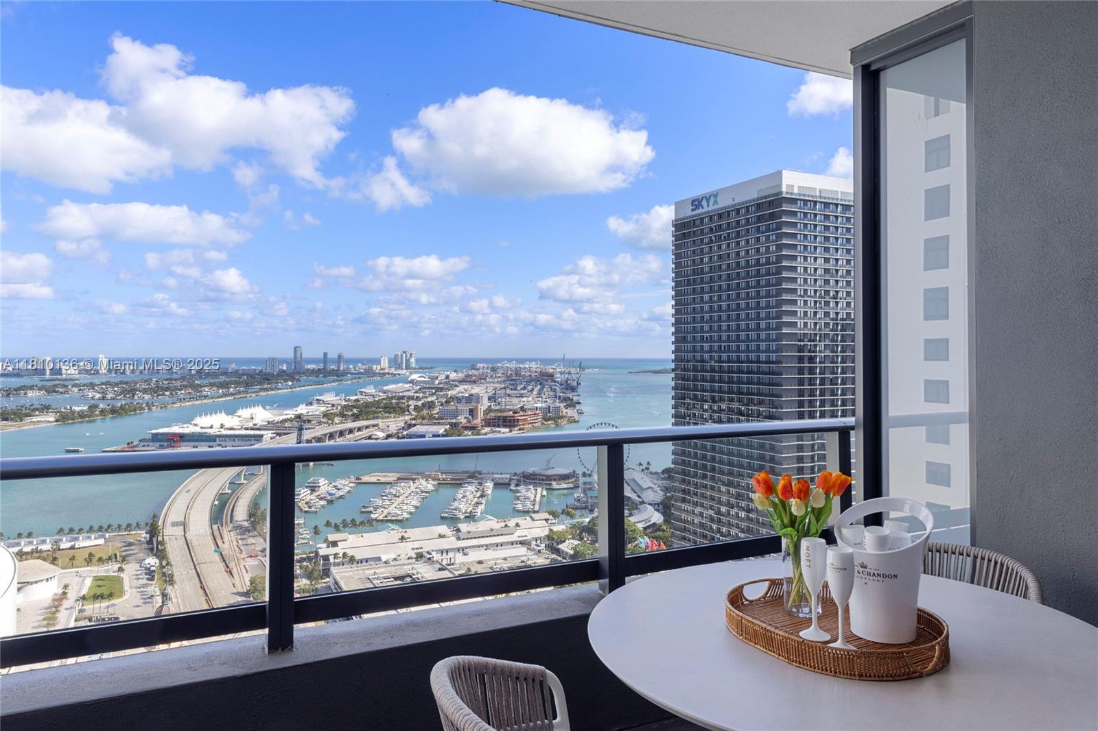A balcony with a round table, champagne bucket, and flowers overlooks a harbor and city — 159 NE 6th St # 3615, Miami FL 33132, MIAMI, FL