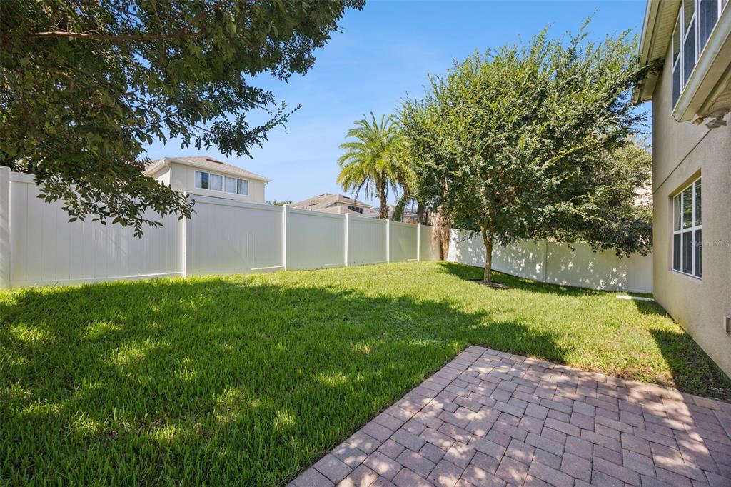A backyard with a paved patio, green lawn, trees, and a white vinyl privacy fence — 13328 OVERSTREET ROAD, WINDERMERE, FL