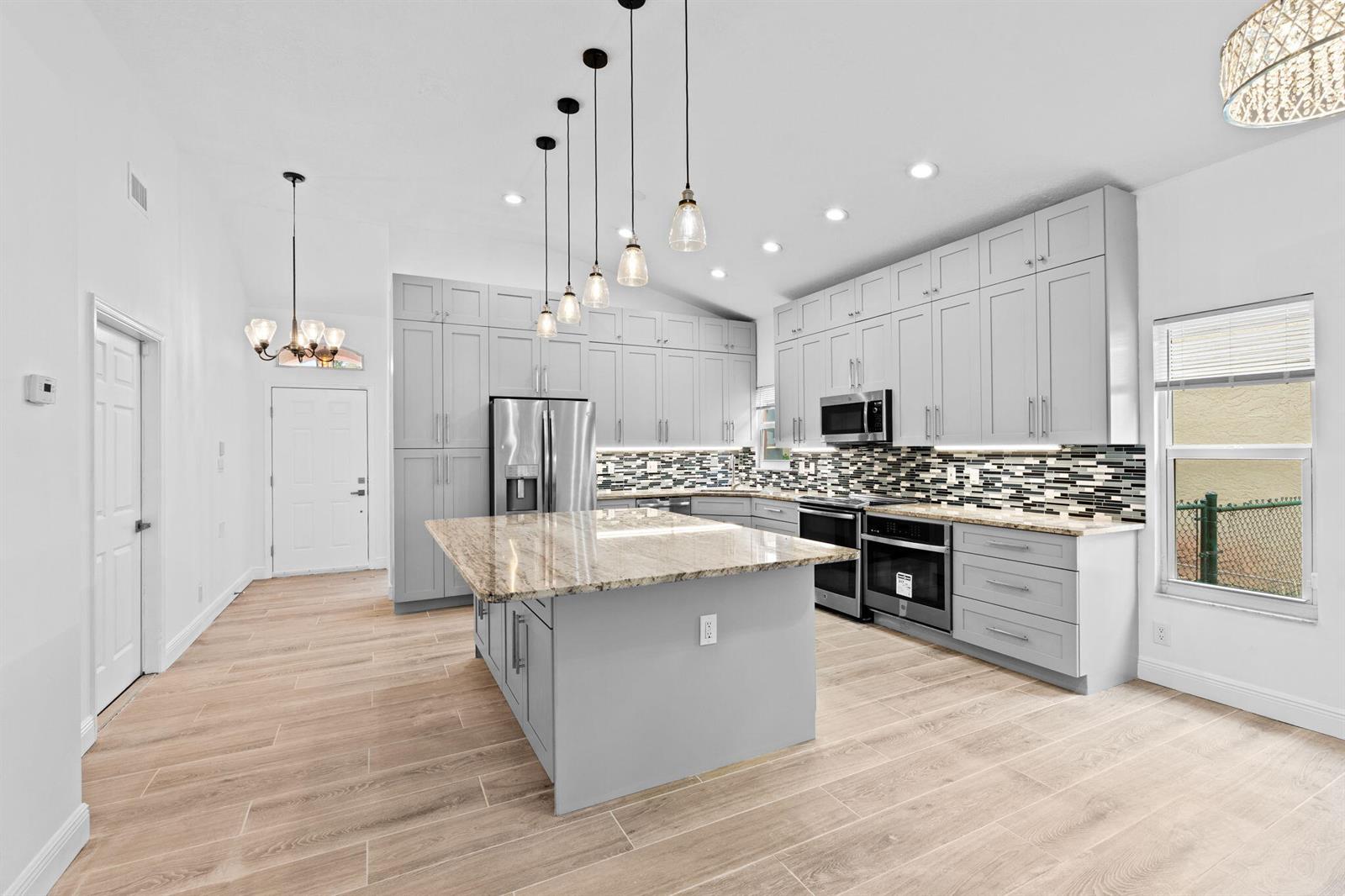 A modern kitchen features gray cabinetry, a granite-topped island, and light wood-look tile flooring — 5122 Elpine Way, Riviera Beach FL 33418, RIVIERA BEACH, FL