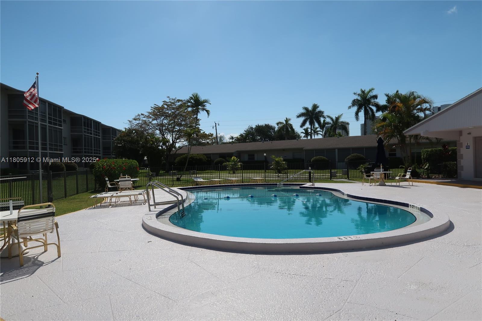 A swimming pool with a white deck is surrounded by palm trees and apartment buildings — 700 Layne Blvd # 112, Hallandale Beach FL 33009, HALLANDALE BEACH, FL
