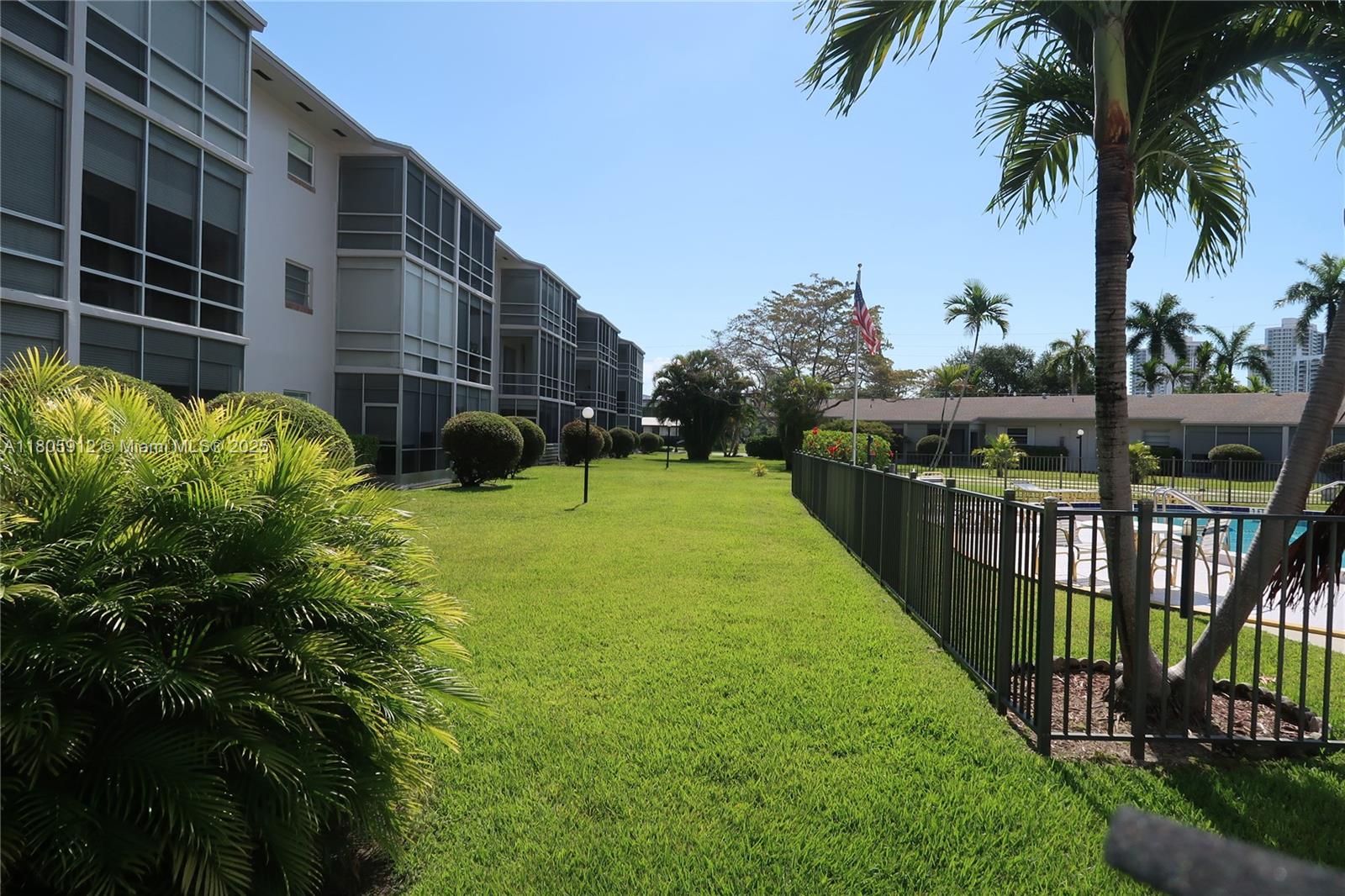 A grassy lawn sits between a multi-story apartment building and a fenced swimming pool area — 700 Layne Blvd # 112, Hallandale Beach FL 33009, HALLANDALE BEACH, FL