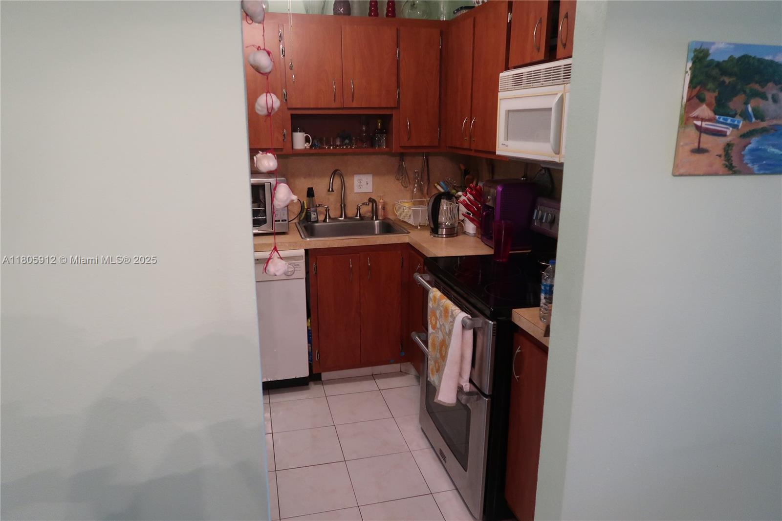 A small galley kitchen features wood cabinets, stainless steel appliances, and white tile flooring — 700 Layne Blvd # 112, Hallandale Beach FL 33009, HALLANDALE BEACH, FL