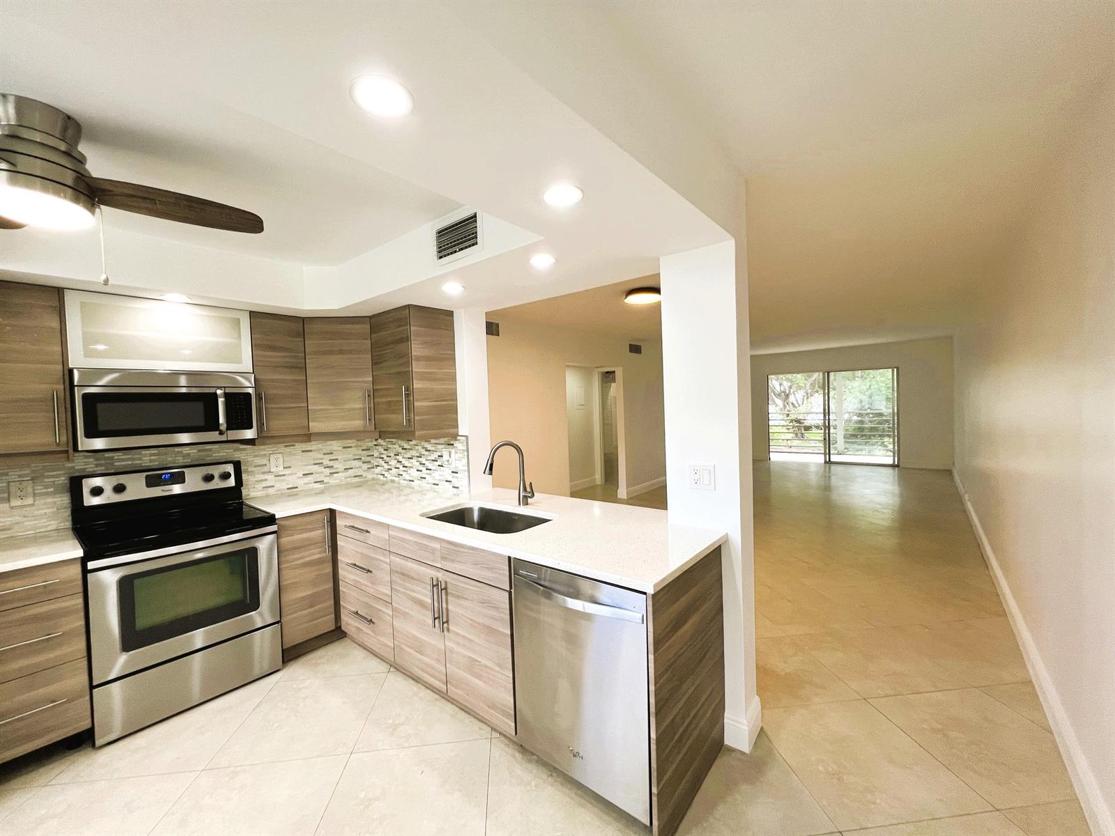 A kitchen with wood-tone cabinets, stainless steel appliances, and quartz countertops opens to a living area — 3001 Portofino Isle # E2, Coconut Creek FL 33066, COCONUT CREEK, FL