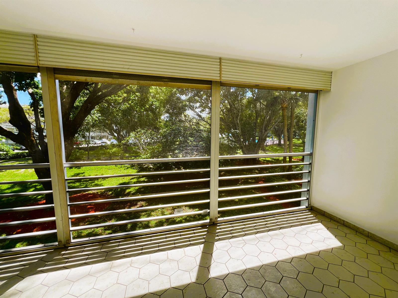 A sunlit enclosed balcony with hexagonal tile flooring and horizontal metal railings overlooking green trees — 3001 Portofino Isle # E2, Coconut Creek FL 33066, COCONUT CREEK, FL