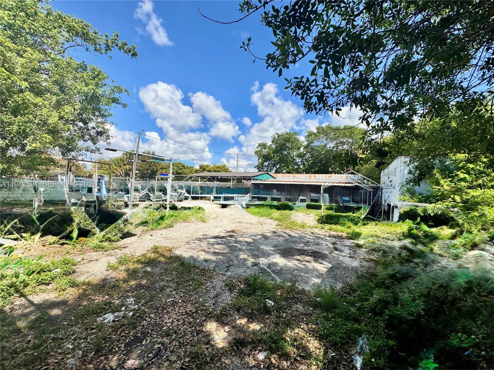 An outdoor view of a gravel lot with overgrown vegetation and a nearby auto shop — 6501 NW 7th Ave, Miami FL 33150, MIAMI, FL