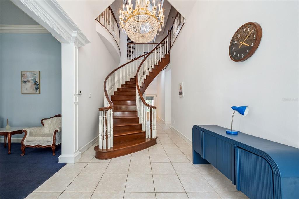 A grand foyer features a curved wooden staircase, a crystal chandelier, and a blue console table — 21033 VIOLET ROAD, BROOKSVILLE, FL