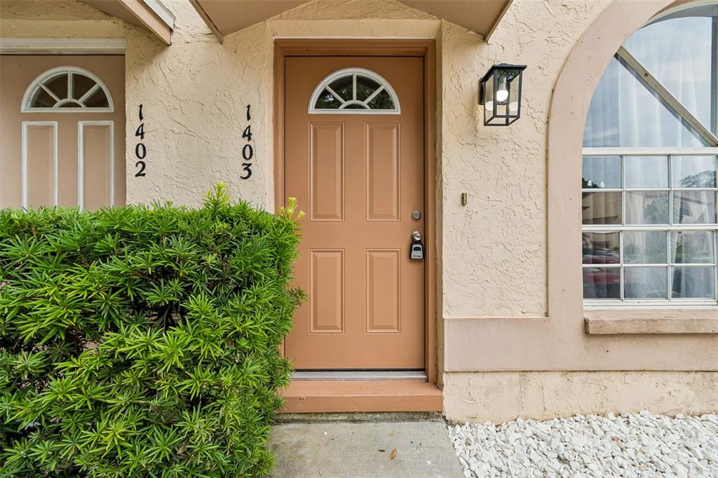 A tan front door with a small arched window sits beside a large window and shrubbery — 1403 BRIGADOON DRIVE, CLEARWATER, FL