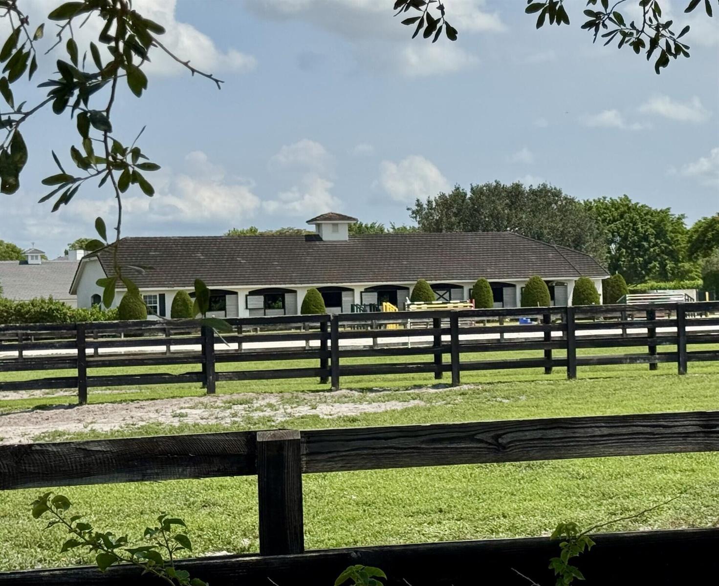 A long, white horse stable with a dark roof sits behind a wooden fence — 14501 Rolling Rock Place, Wellington FL 33414, WELLINGTON, FL