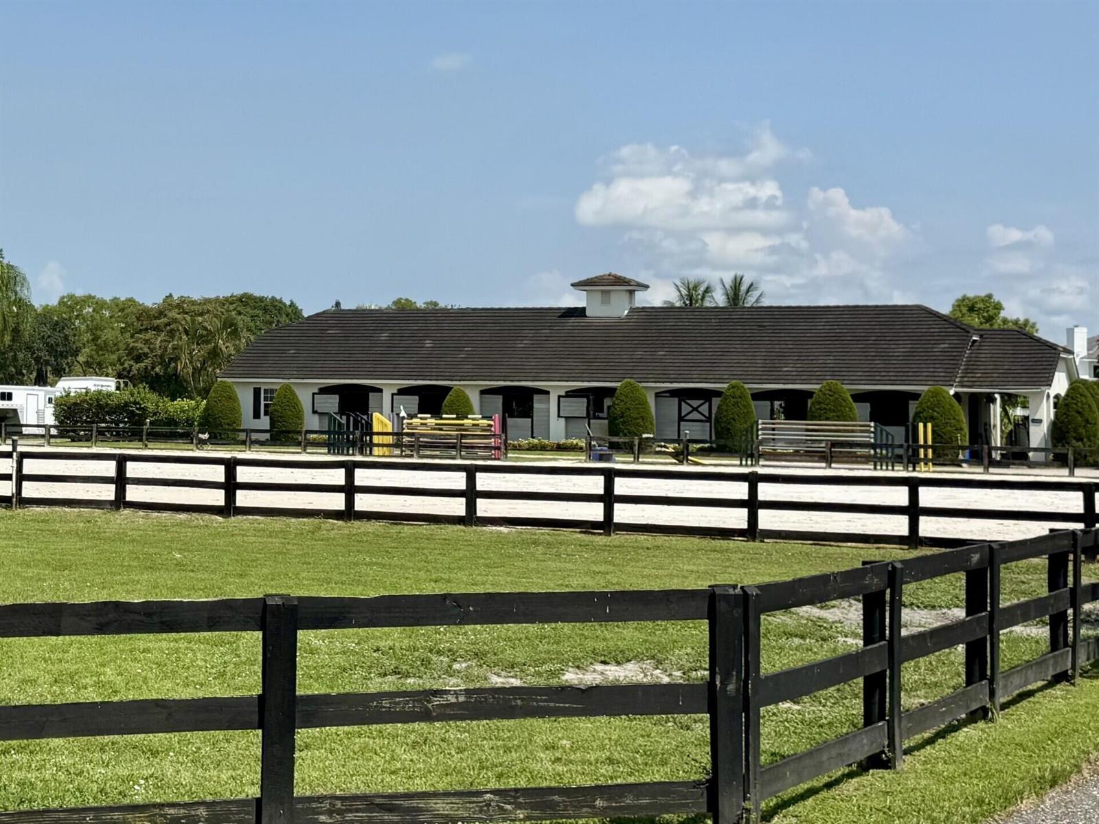 A white horse stable with a dark roof sits behind a black wooden fence — 14501 Rolling Rock Place, Wellington FL 33414, WELLINGTON, FL