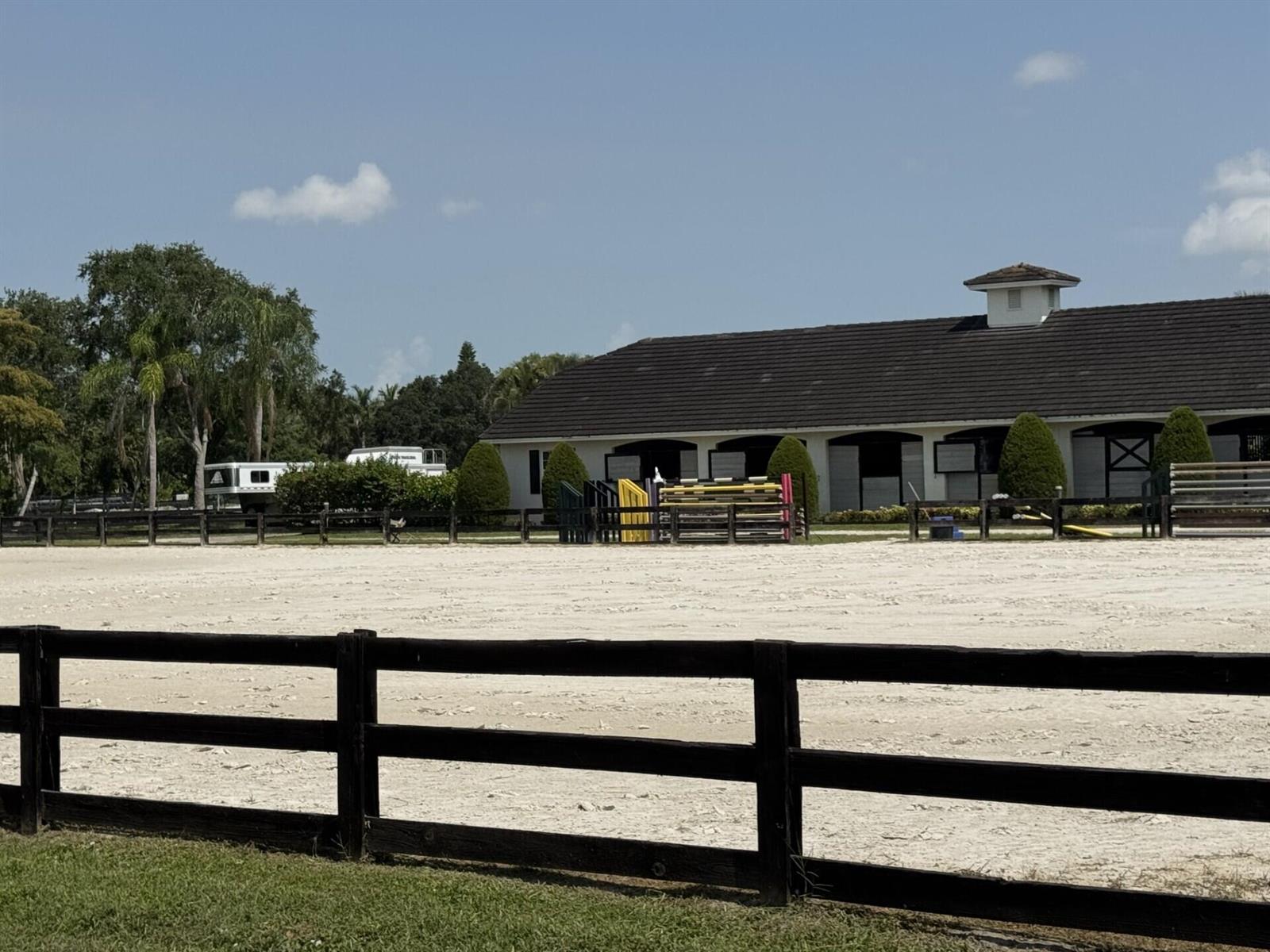 A white stable building sits behind a large sand riding arena with a wooden fence — 14501 Rolling Rock Place, Wellington FL 33414, WELLINGTON, FL