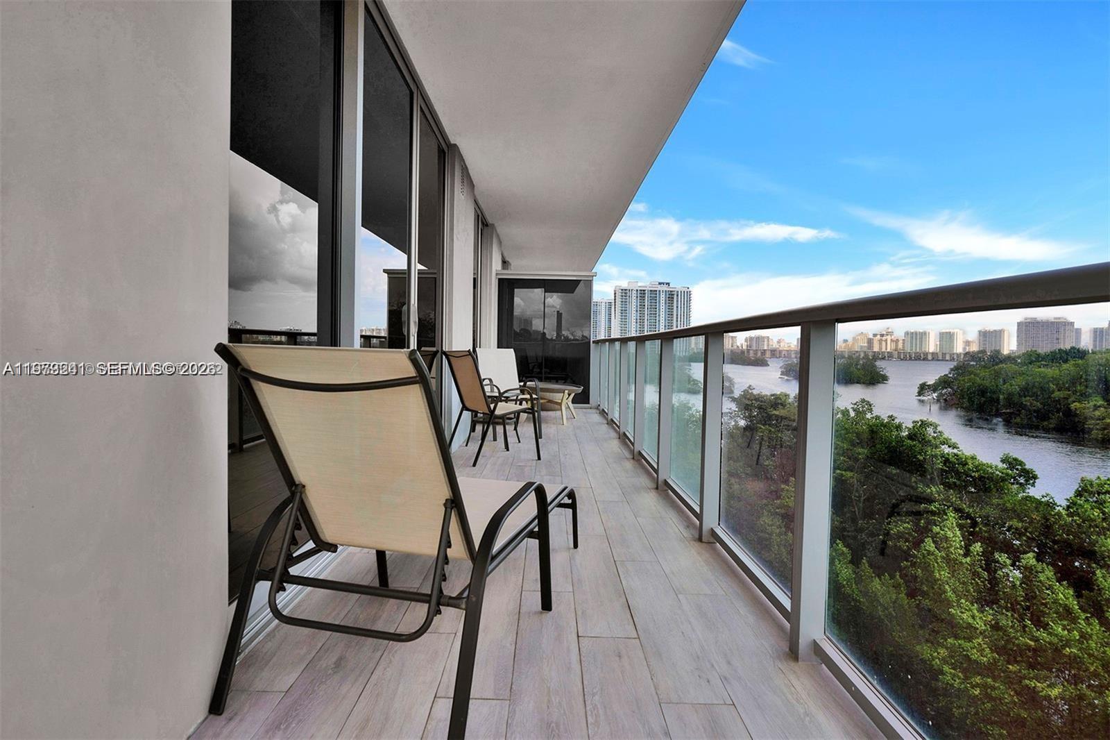 A balcony features light wood-look tile flooring, glass railings, and outdoor chairs overlooking water — 16385 Biscayne Blvd # 421, North Miami Beach FL 33160, NORTH MIAMI BEACH, FL
