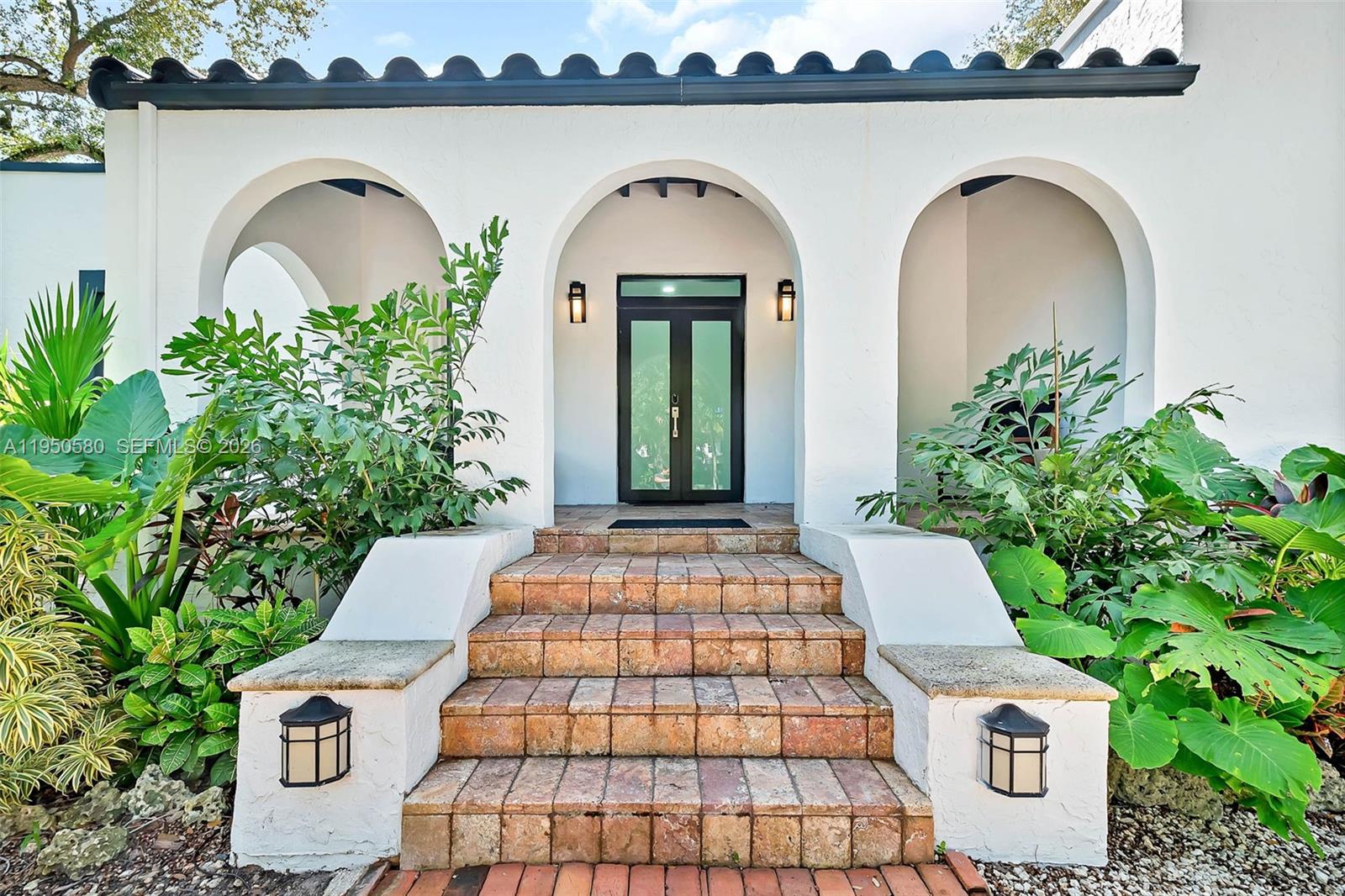 A white stucco house exterior features a central arched doorway with brick steps and landscaping — 1702 Espanola Dr, Miami FL 33133, MIAMI, FL
