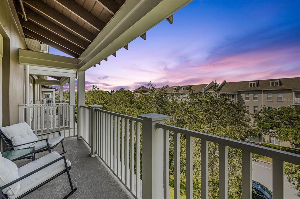 A covered balcony with white railings and two chairs overlooks trees and a neighboring building — 1410 CELEBRATION AVENUE UNIT 407, CELEBRATION, FL