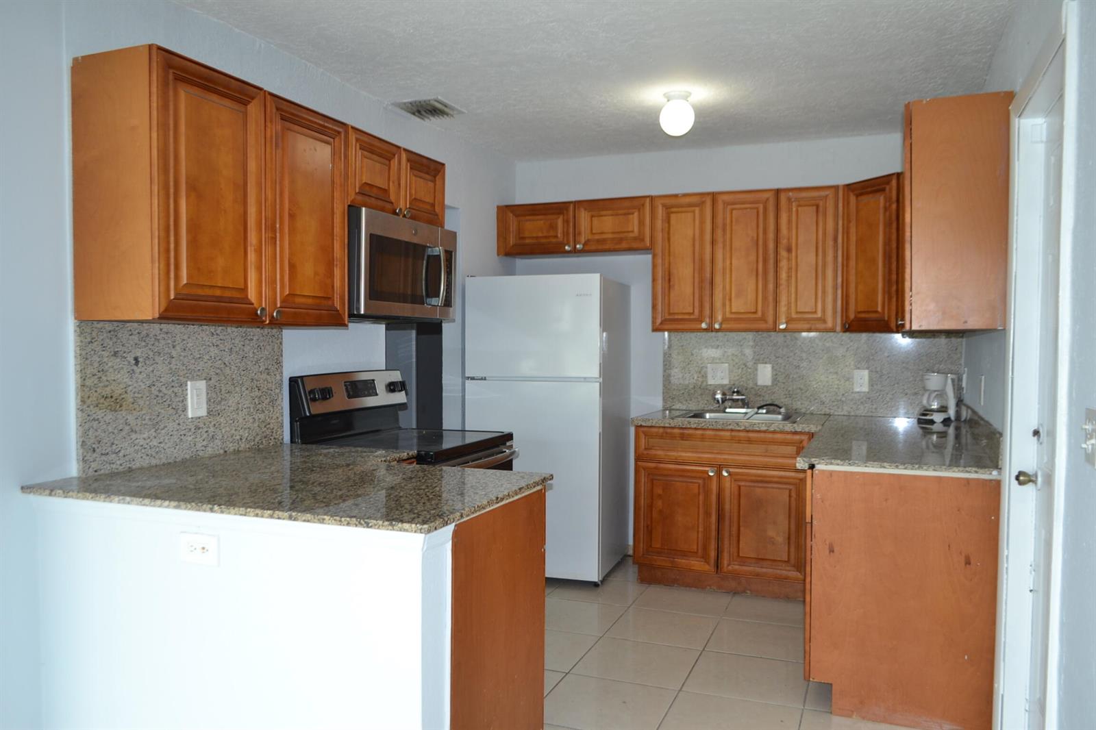 A kitchen features wood cabinets, granite countertops, a white refrigerator, and a stainless steel microwave — 608 NW 20th Court, Pompano Beach FL 33060, POMPANO BEACH, FL