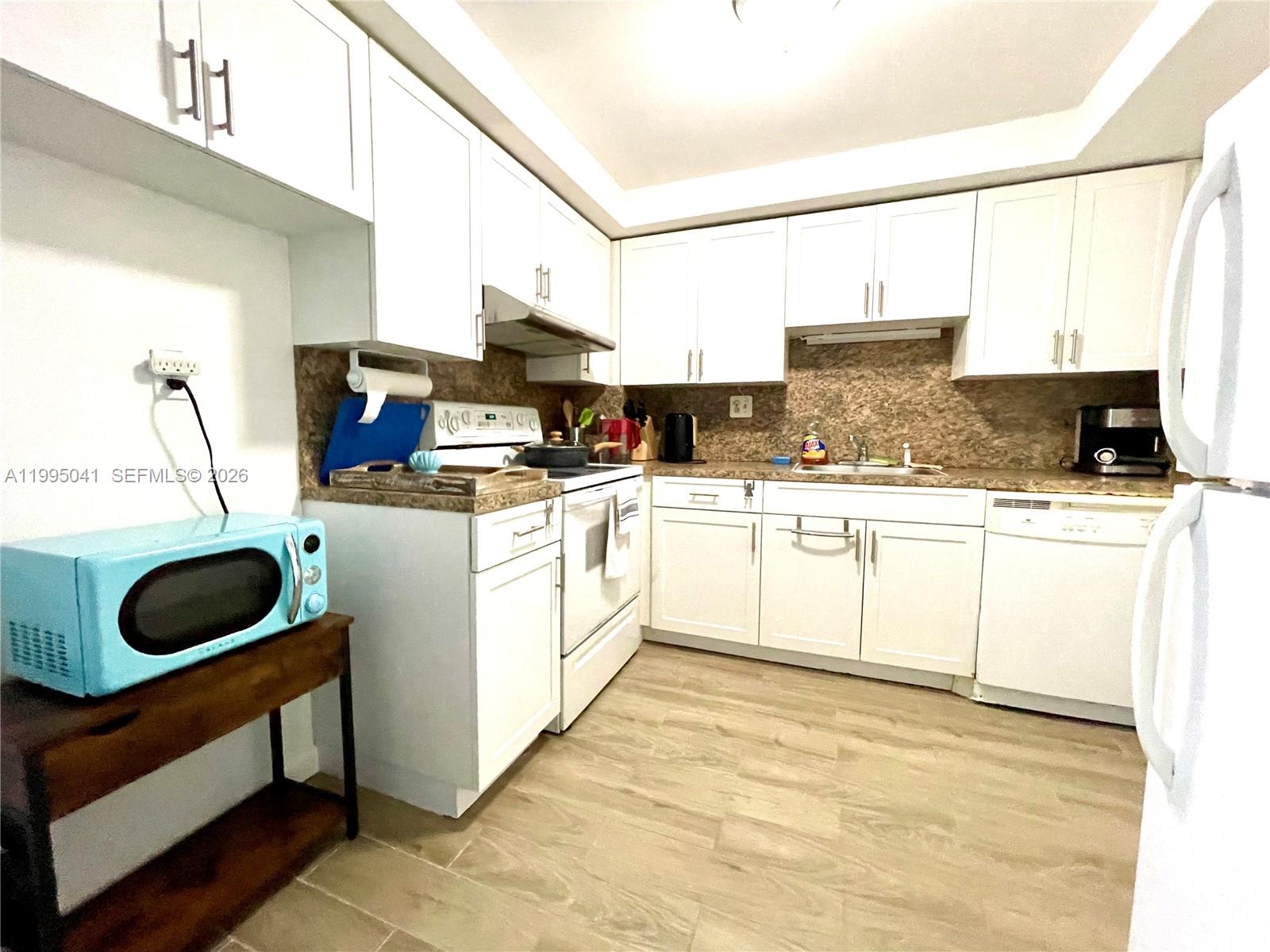 A kitchen features white cabinets, light wood-look flooring, and a light blue microwave — 1913 S OCEAN DR # 129, Hallandale Beach FL 33009, HALLANDALE BEACH, FL