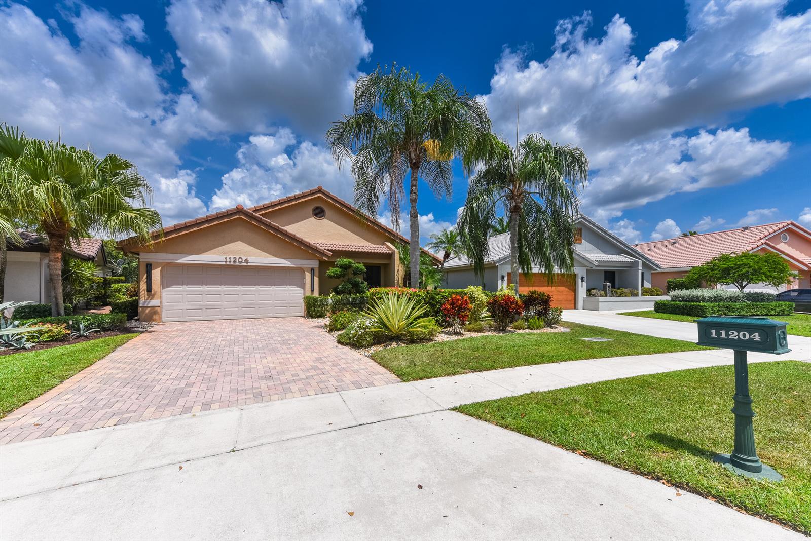 A tan house with a paved driveway and palm trees sits under a cloudy sky — 11204 Cloverleaf Circle, Boca Raton FL 33428, BOCA RATON, FL