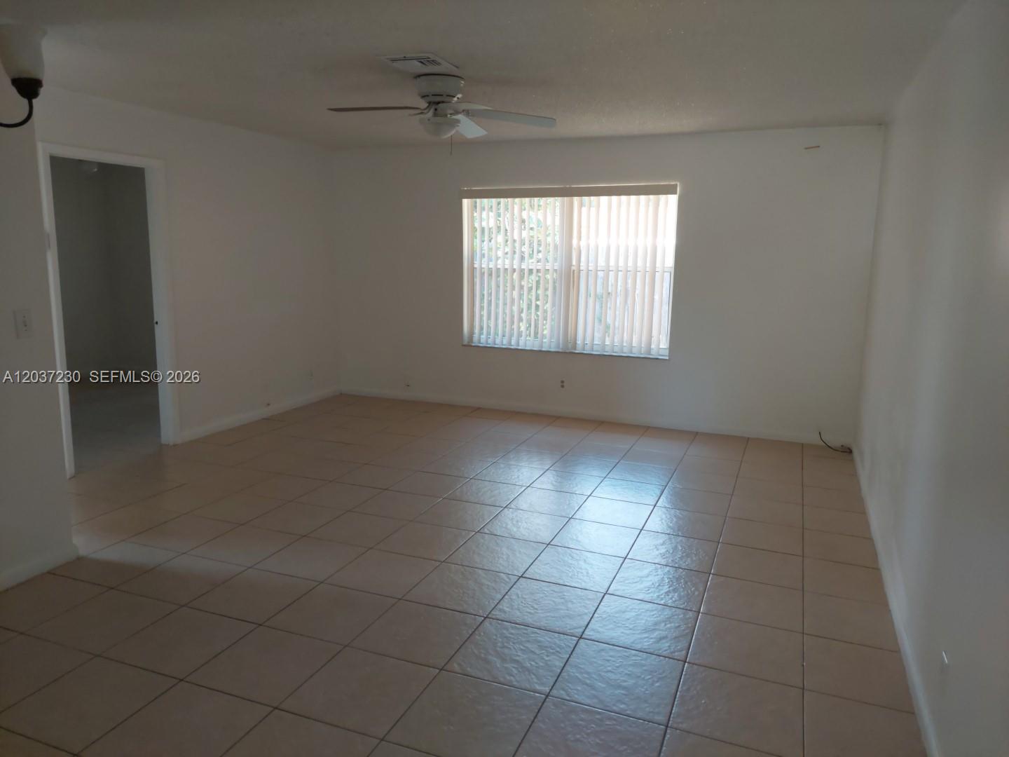 An empty room features white walls, tan tile flooring, a ceiling fan, and vertical blinds — 2319 Johnson St # 1, Hollywood FL 33020, HOLLYWOOD, FL