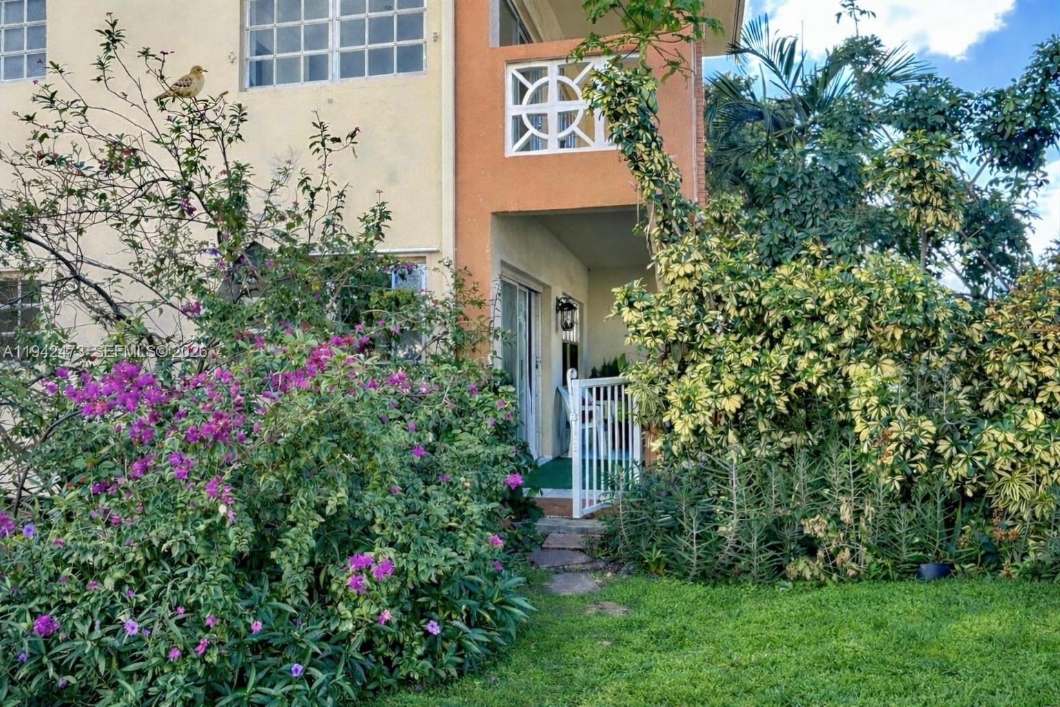 A ground-level entrance to a building features a white gate, lush landscaping, and purple flowers — 11840 NE 19th Dr # 9, North Miami FL 33181, NORTH MIAMI, FL