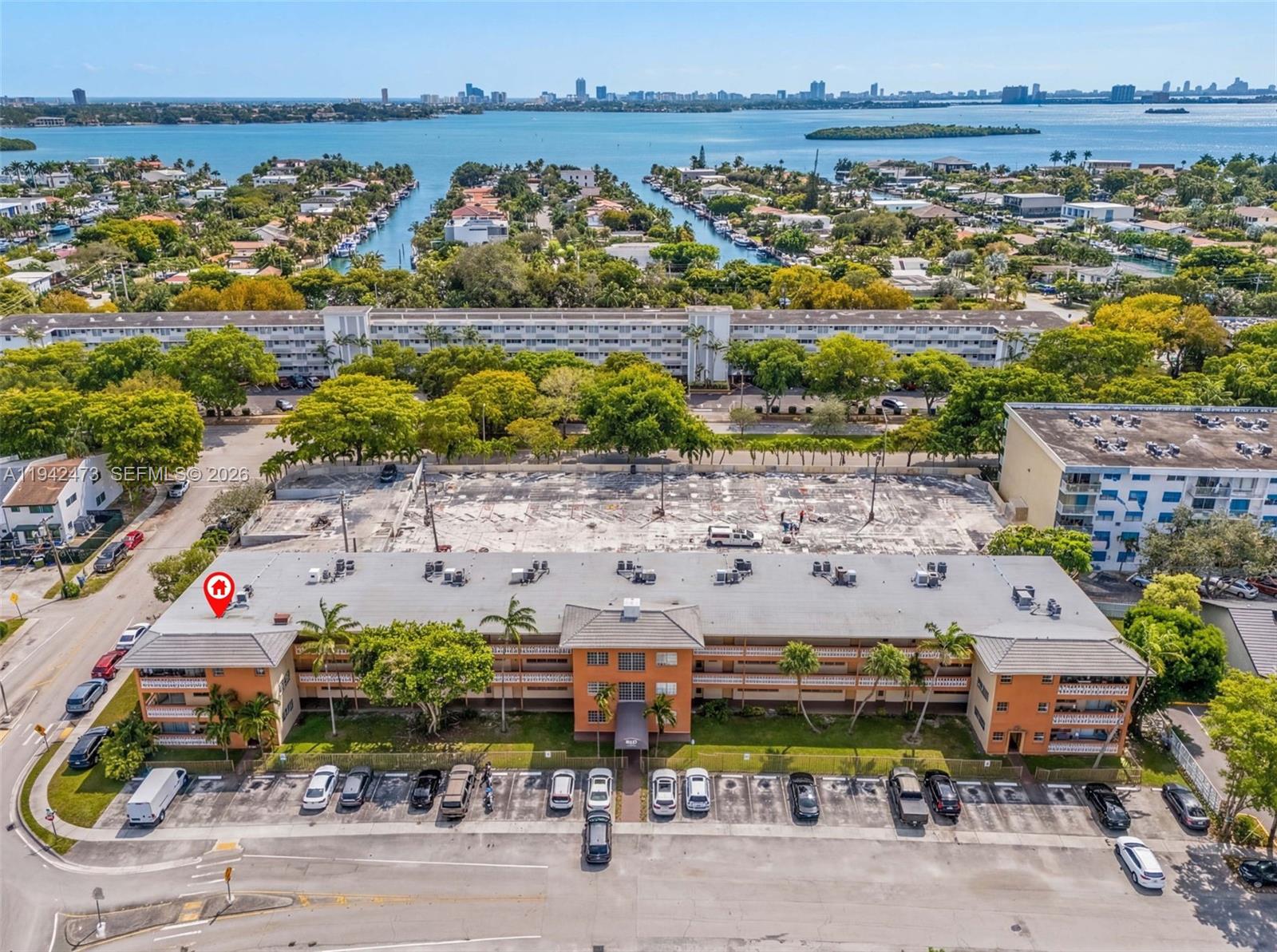 An aerial view shows a multi-story orange apartment building with a parking lot and waterfront surroundings — 11840 NE 19th Dr # 9, North Miami FL 33181, NORTH MIAMI, FL