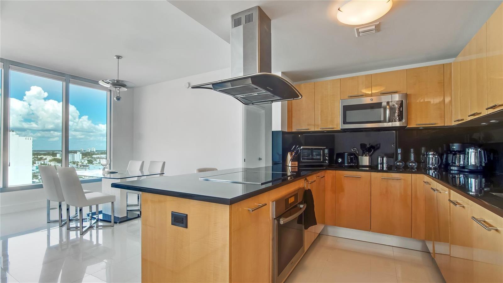 A modern kitchen features light wood cabinets, black countertops, stainless steel appliances, and a dining area — 6899 Collins Avenue # 1203, Miami Beach FL 33141, MIAMI BEACH, FL