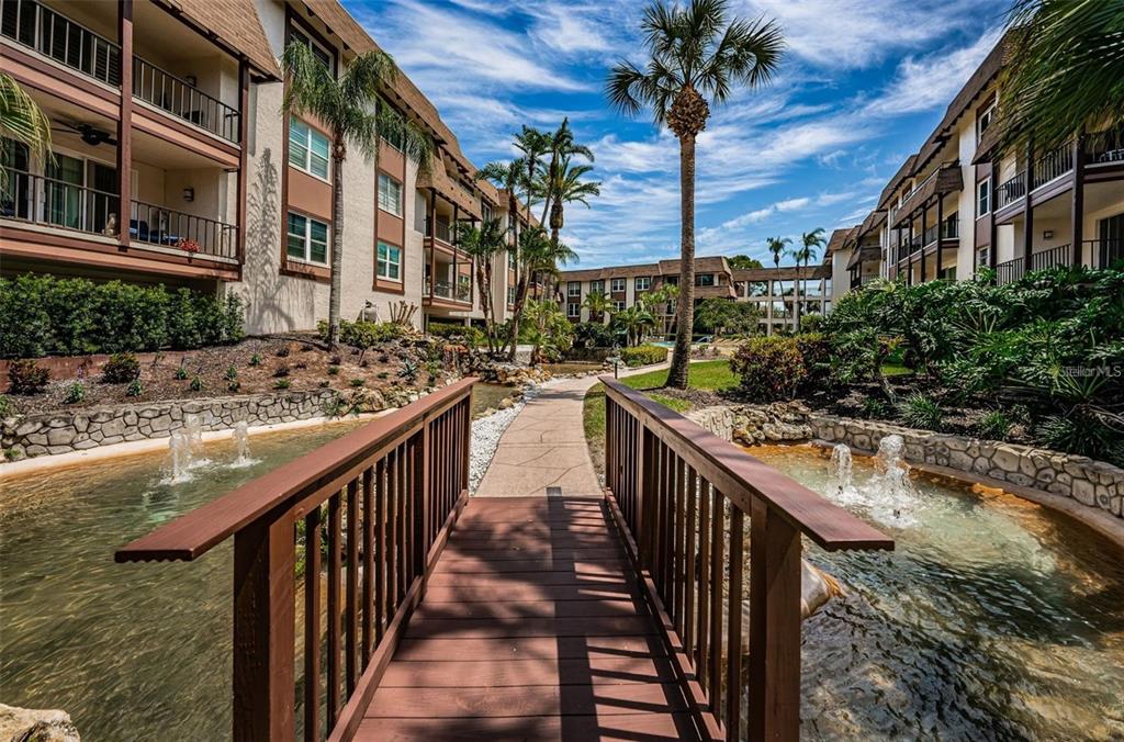 A wooden footbridge crosses a pond with fountains between two multi-story apartment buildings — 3021 COUNTRYSIDE BOULEVARD UNIT 44A, CLEARWATER, FL