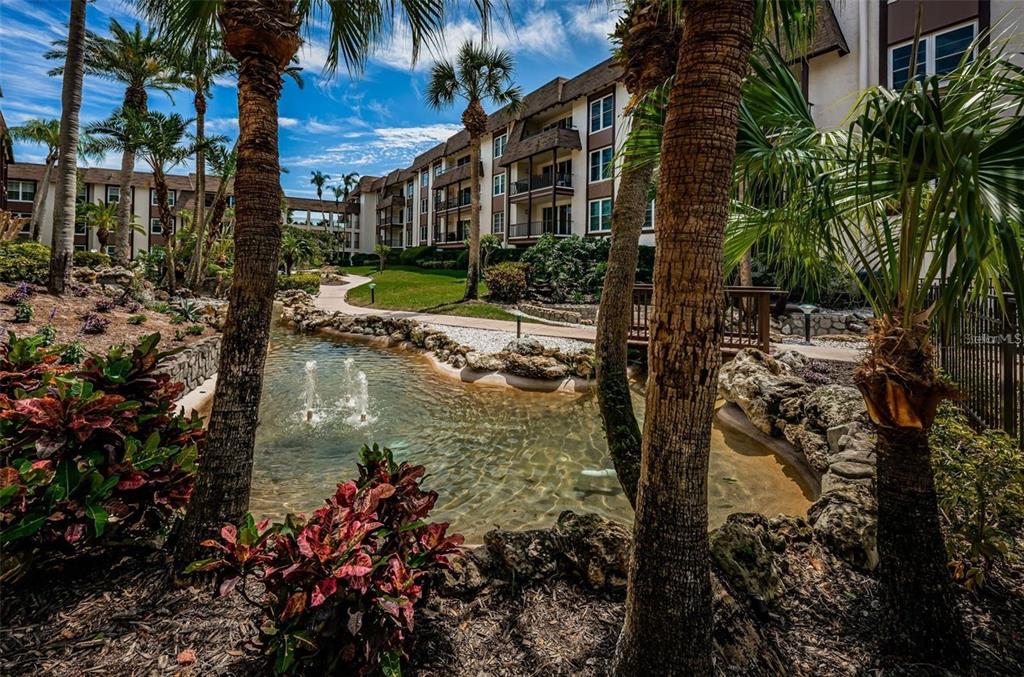 A landscaped courtyard features a small pond with fountains, palm trees, and apartment buildings — 3021 COUNTRYSIDE BOULEVARD UNIT 44A, CLEARWATER, FL