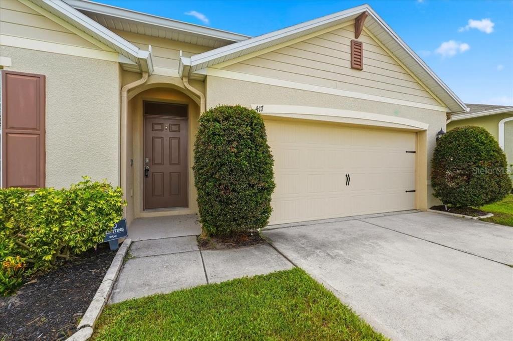 The exterior of a single-story house features a tan garage, brown front door, and landscaping — 417 CORSO LOOP, WINTER HAVEN, FL