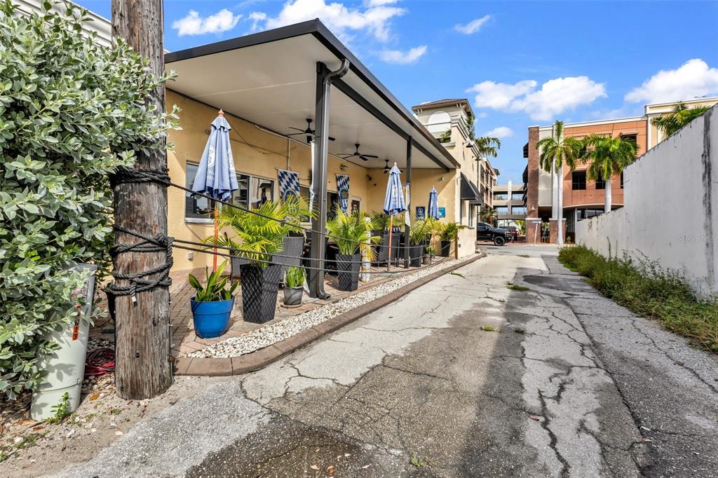 An outdoor patio area with tables, umbrellas, and potted plants sits beside a paved alleyway — 111 W OLYMPIA AVENUE, PUNTA GORDA, FL