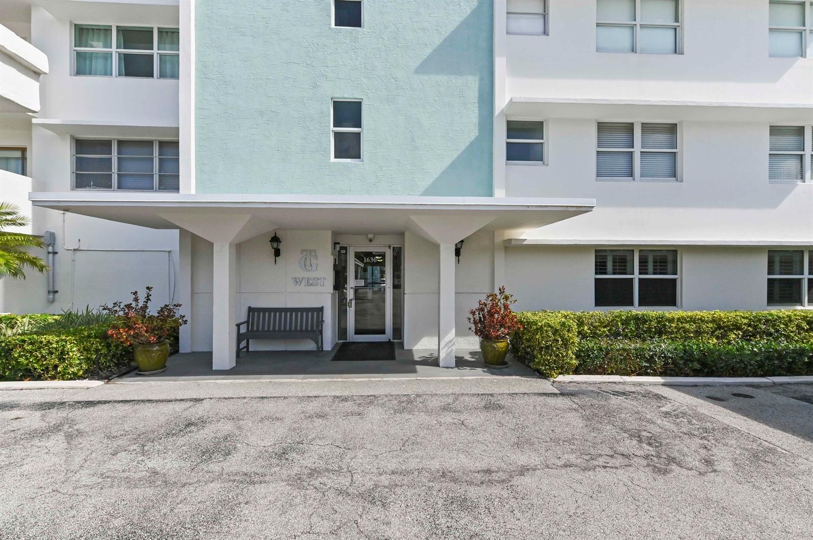 The exterior of a multi-story building features a covered entrance with a bench and landscaping — 1630 N Ocean Boulevard # 215, Pompano Beach FL 33062, POMPANO BEACH, FL