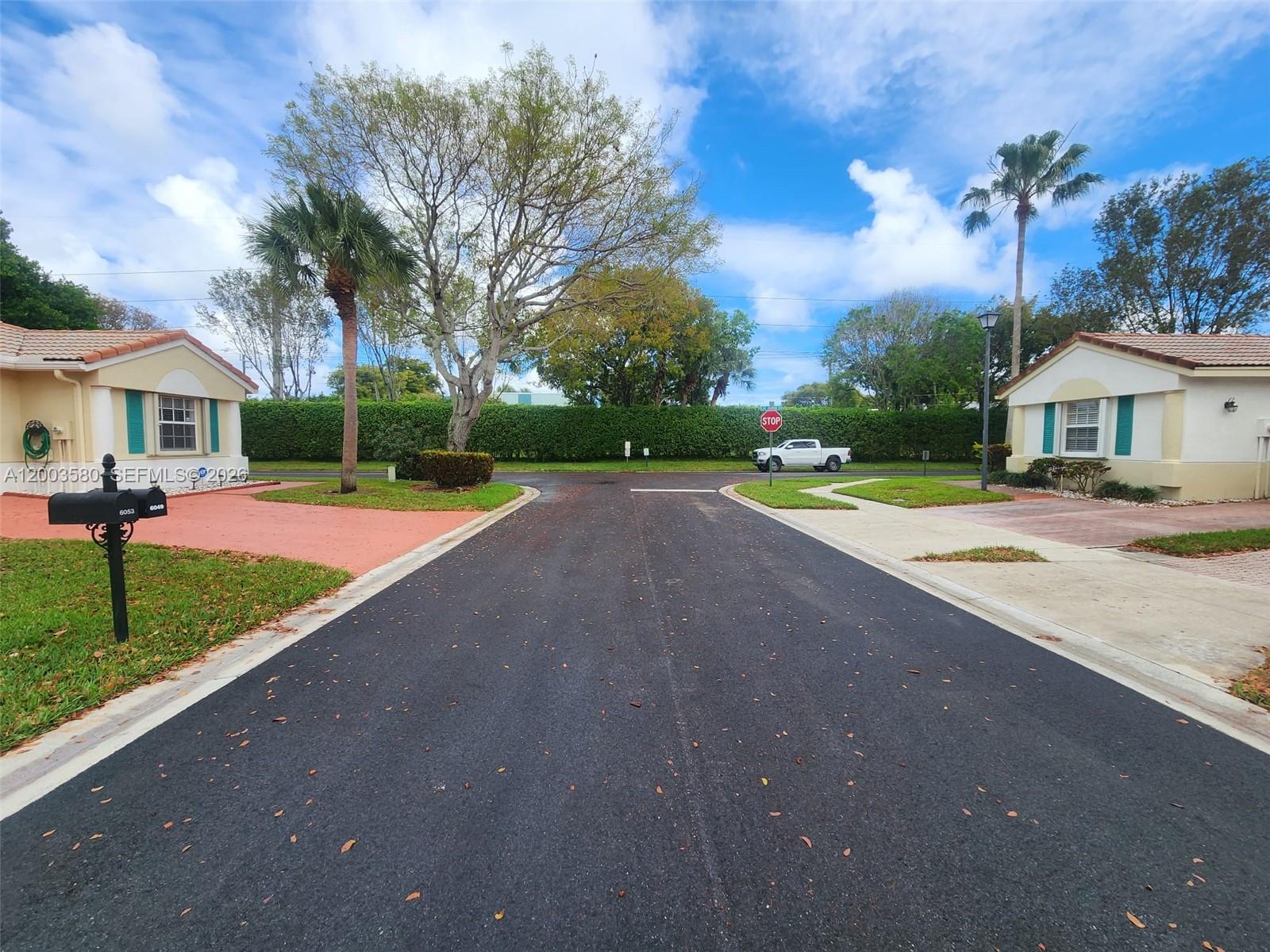 A paved street leads to a T-intersection between two single-story houses with palm trees — 6053 Heliconia Rd, Delray Beach FL 33484, DELRAY BEACH, FL