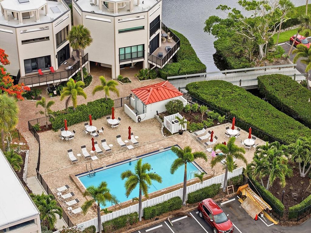 An aerial view shows a swimming pool with patio seating, palm trees, and nearby buildings — 3705 E BAY DRIVE UNIT 117, HOLMES BEACH, FL