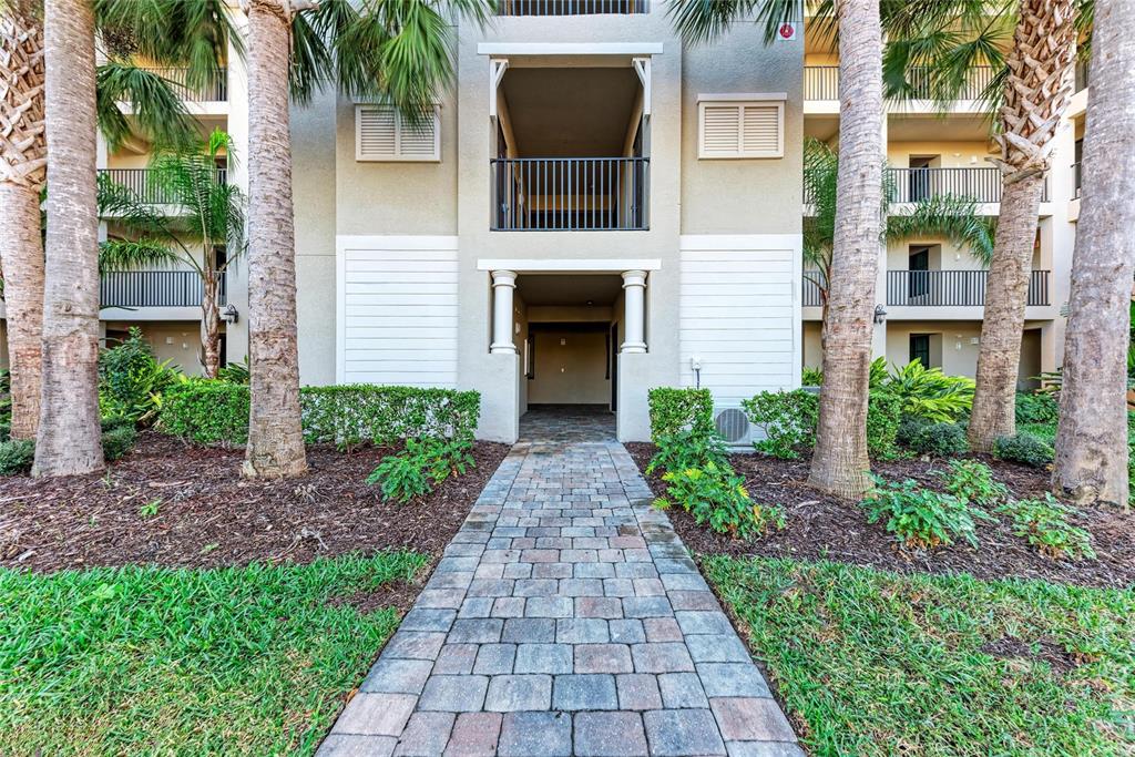 A stone walkway leads to the entrance of a multi-story apartment building with palm trees — 18114 GAWTHROP DRIVE UNIT 204, BRADENTON, FL