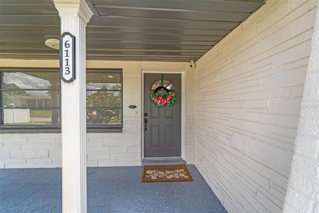 A gray front door with a floral wreath sits under a covered porch with brick walls — 6113 9TH AVENUE, NEW PORT RICHEY, FL