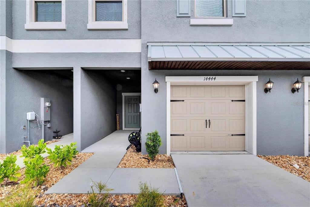 A gray townhome exterior features a beige garage door, a covered entryway, and landscaping — 14444 EDITORS NOTE STREET, SUN CITY CENTER, FL