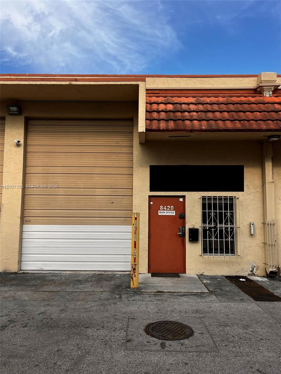 A beige industrial building exterior features a roll-up garage door and a red office door — 8420 NW 61st St, Miami FL 33166, MIAMI, FL