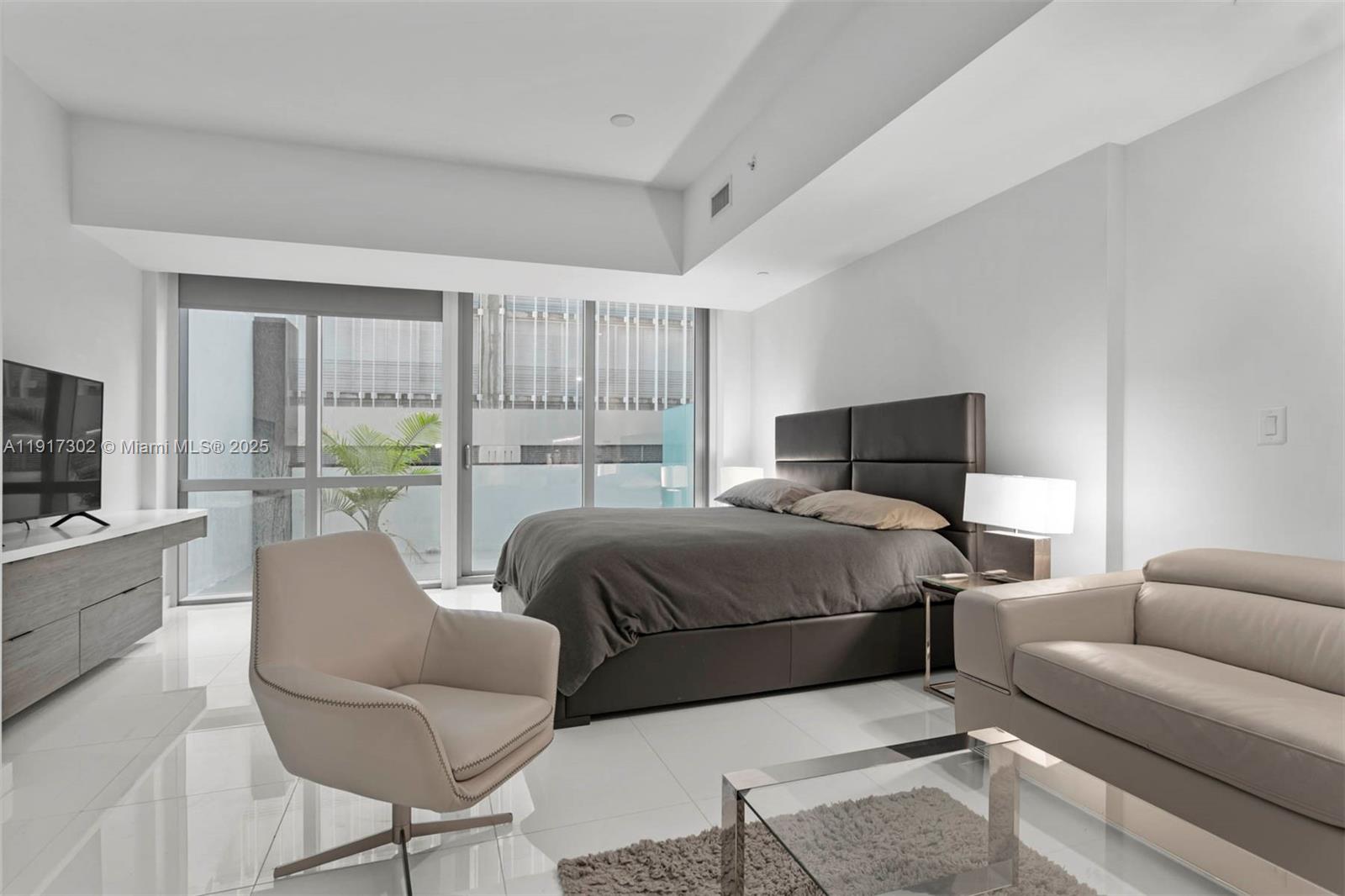 A modern bedroom features a dark upholstered bed, light leather seating, and floor-to-ceiling windows — 851 NE 1st Ave # 300, Miami FL 33132, MIAMI, FL