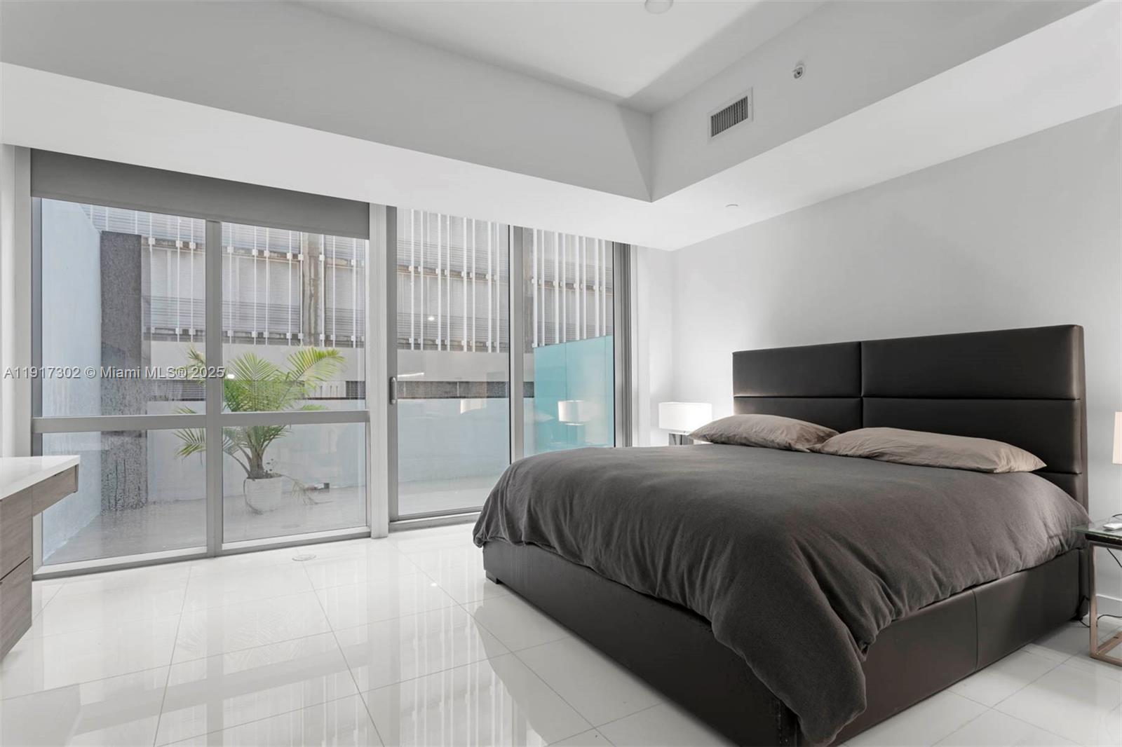 A modern bedroom features a dark upholstered bed, white tile floors, and floor-to-ceiling windows — 851 NE 1st Ave # 300, Miami FL 33132, MIAMI, FL
