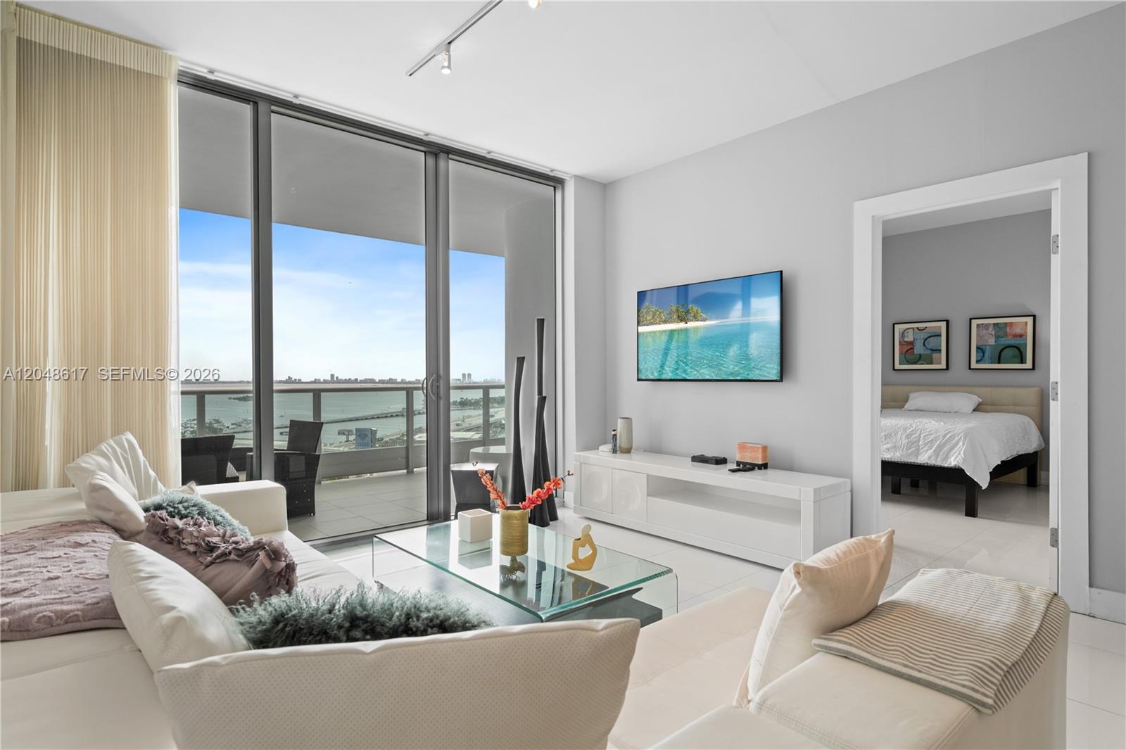 A modern living room features a white sectional, glass coffee table, and balcony with water views — 900 Biscayne Blvd # 2408, Miami FL 33132, MIAMI, FL