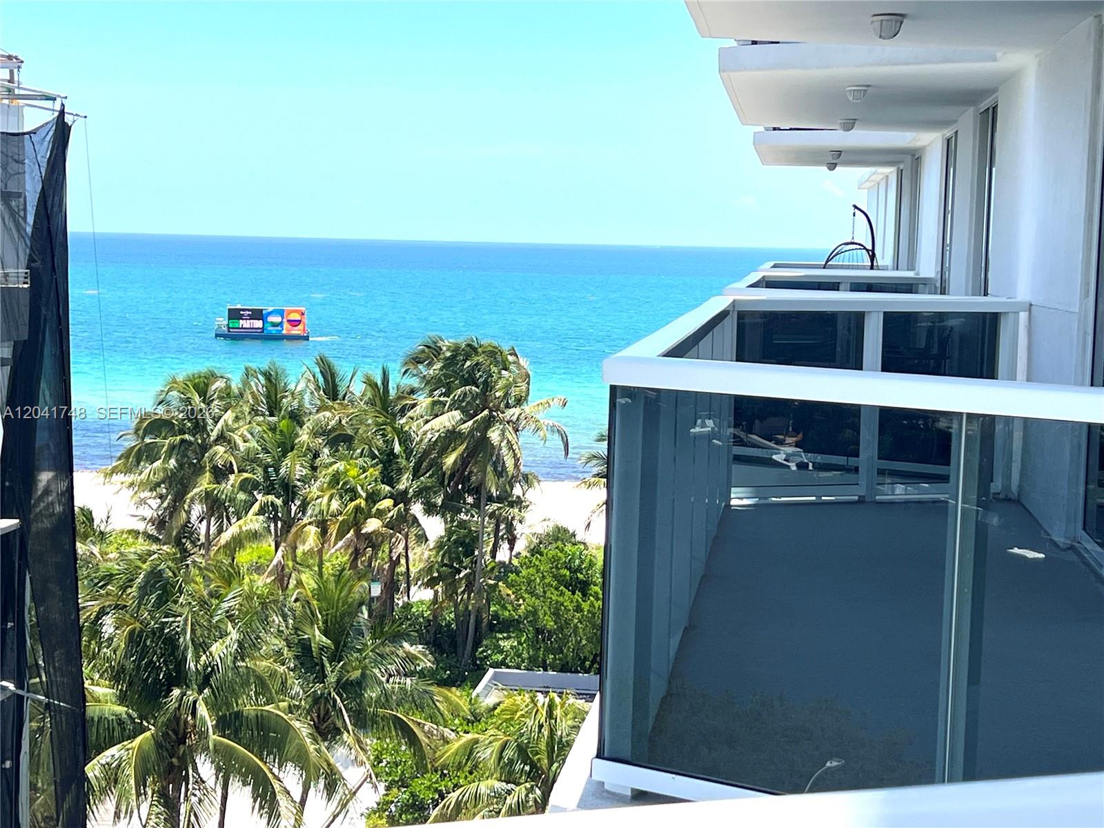 A balcony with glass railings overlooks palm trees, a sandy beach, and the ocean — 2401 Collins Ave # 1009, Miami Beach FL 33140, MIAMI BEACH, FL