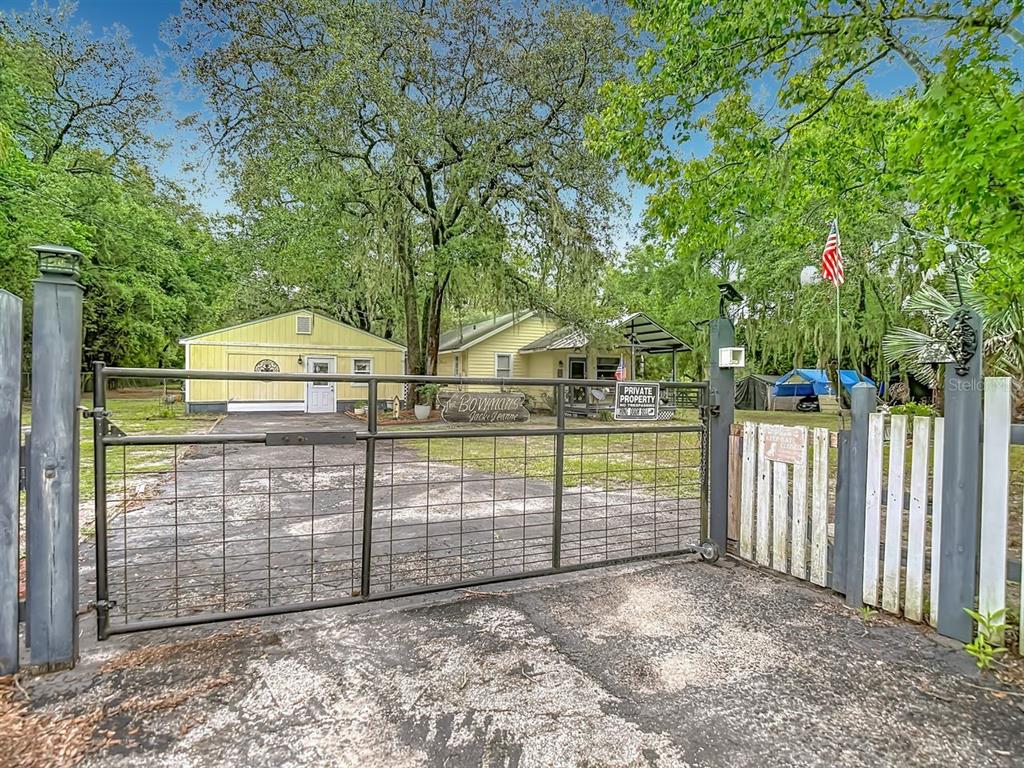 A metal gate opens to a driveway leading toward a yellow house and detached garage — 11705 MARY LOU DRIVE, HUDSON, FL