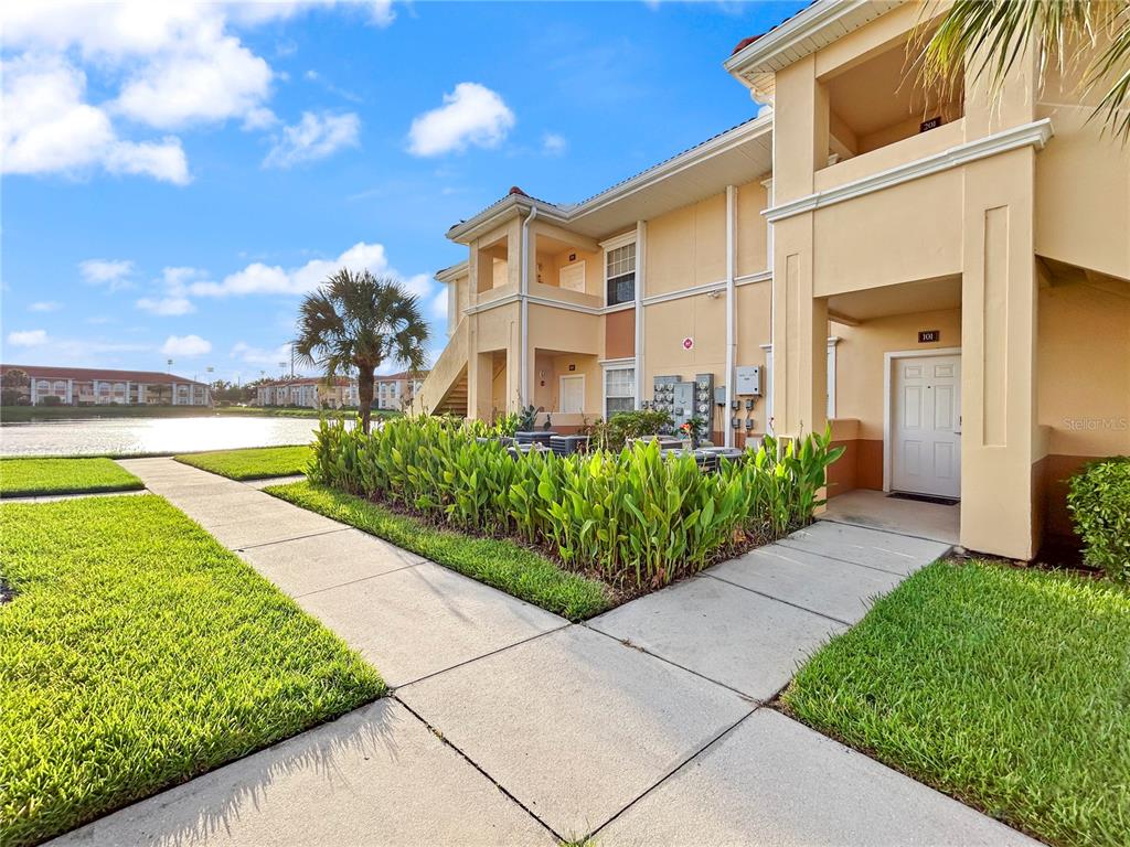 A two-story apartment building exterior features a concrete walkway, landscaping, and a nearby pond — 1025 VILLAGIO CIRCLE UNIT 101, SARASOTA, FL