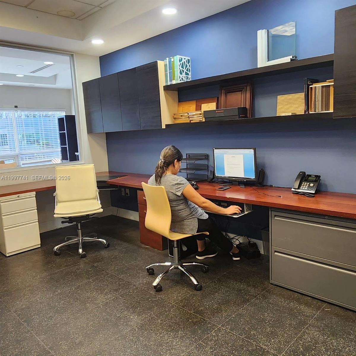 An office features a long wooden desk, dark cabinetry, open shelving, and a person working — 2441 NW 93 # B, Doral FL 33172, DORAL, FL