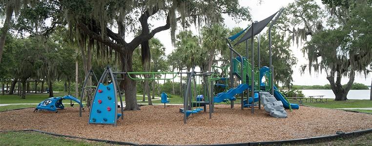 A playground with a climbing wall and slides sits on a wood-chipped surface near trees — 3394 HIGHLANDS ROAD, PUNTA GORDA, FL