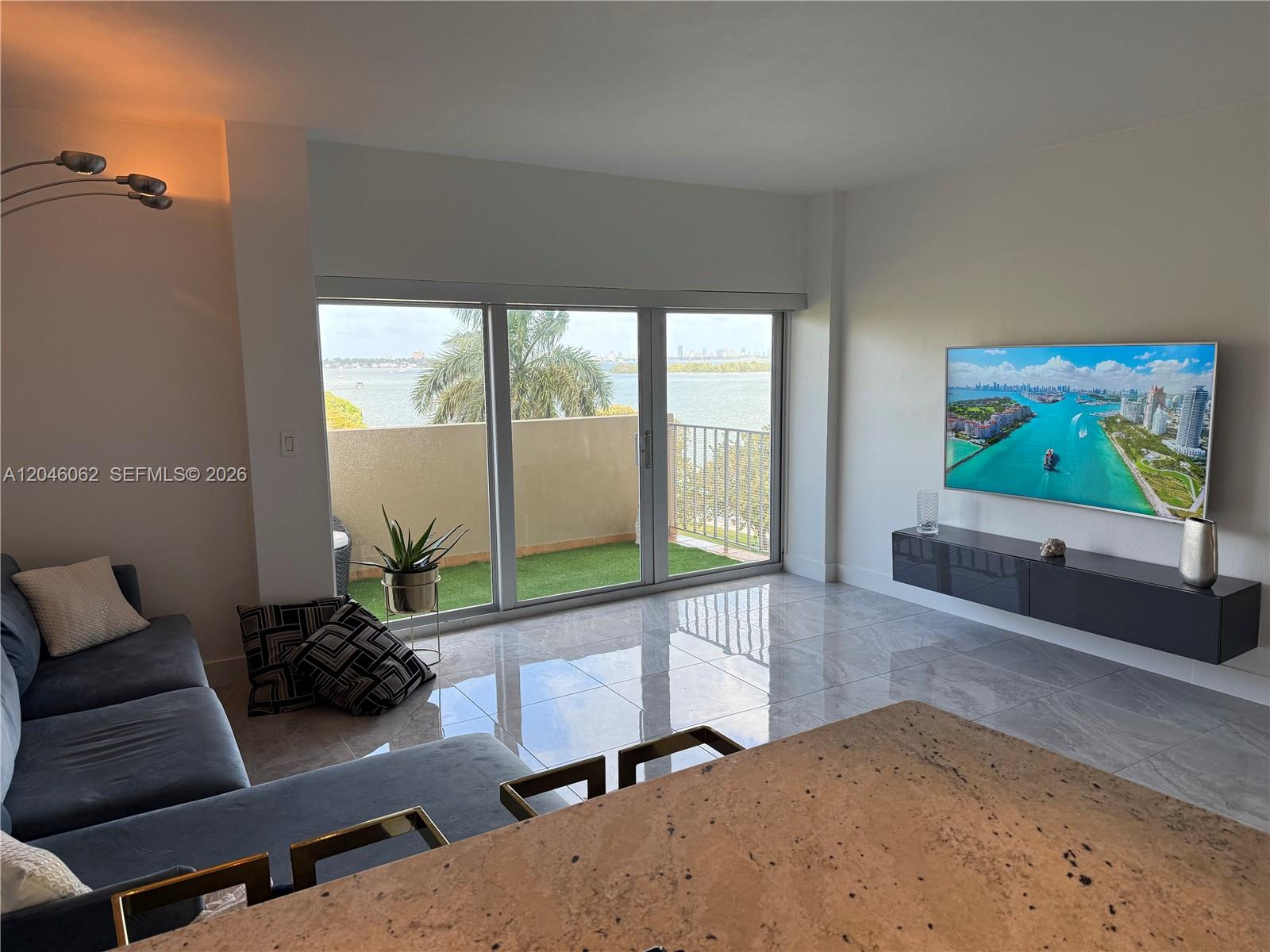 A living room features a gray sectional, a wall-mounted television, and a balcony overlooking water — 880 NE 69th St # 5Q, Miami FL 33138, MIAMI, FL