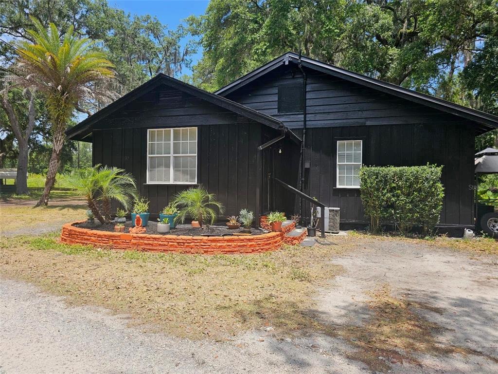 A black-painted house features a curved orange brick planter box filled with potted plants — 19240 SE HIGHWAY 42, WEIRSDALE, FL
