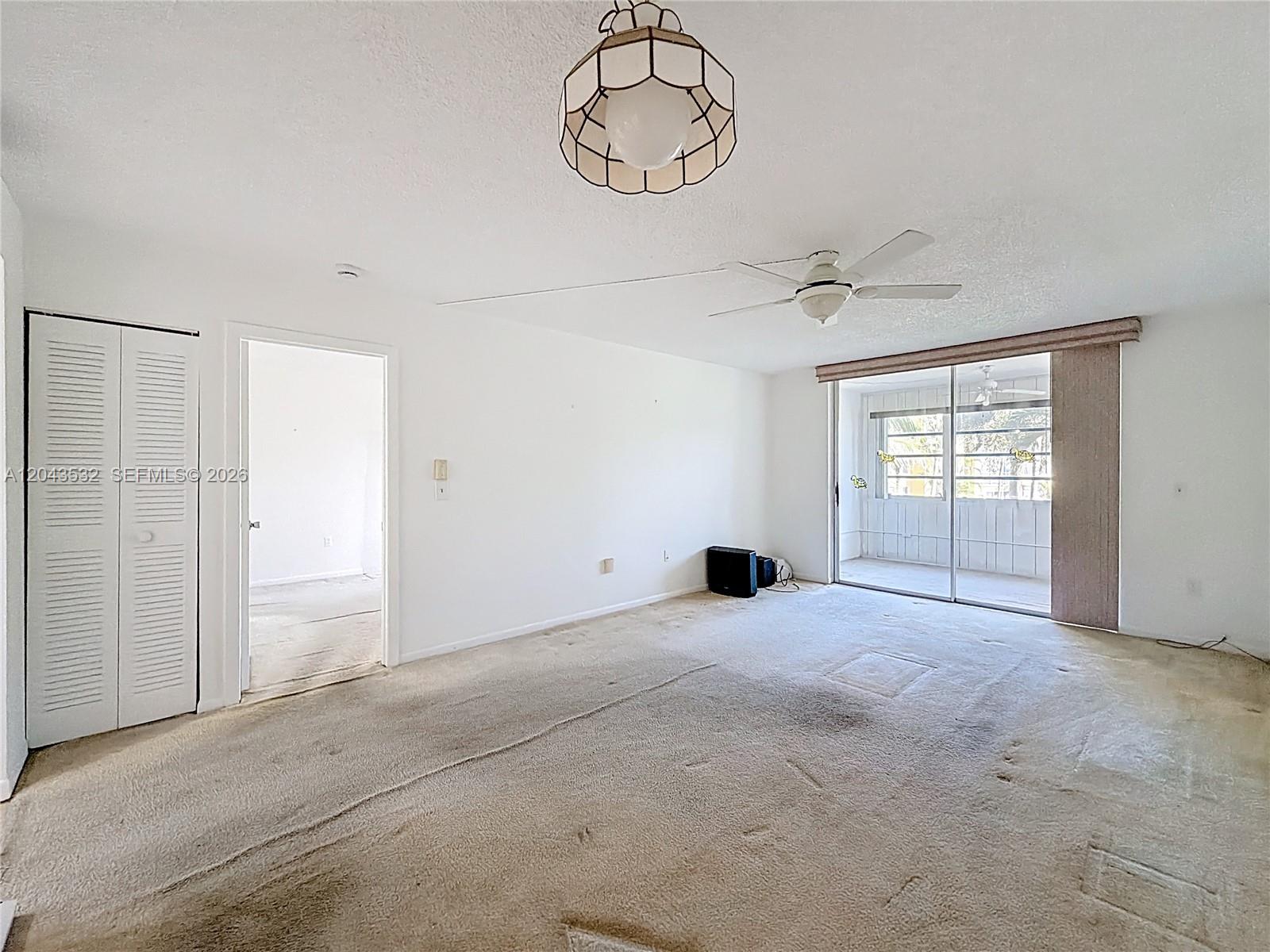An empty living room features beige carpeting, white walls, a ceiling fan, and sliding glass doors — 6750 Royal Palm Blvd # 209E, Margate FL 33063, MARGATE, FL