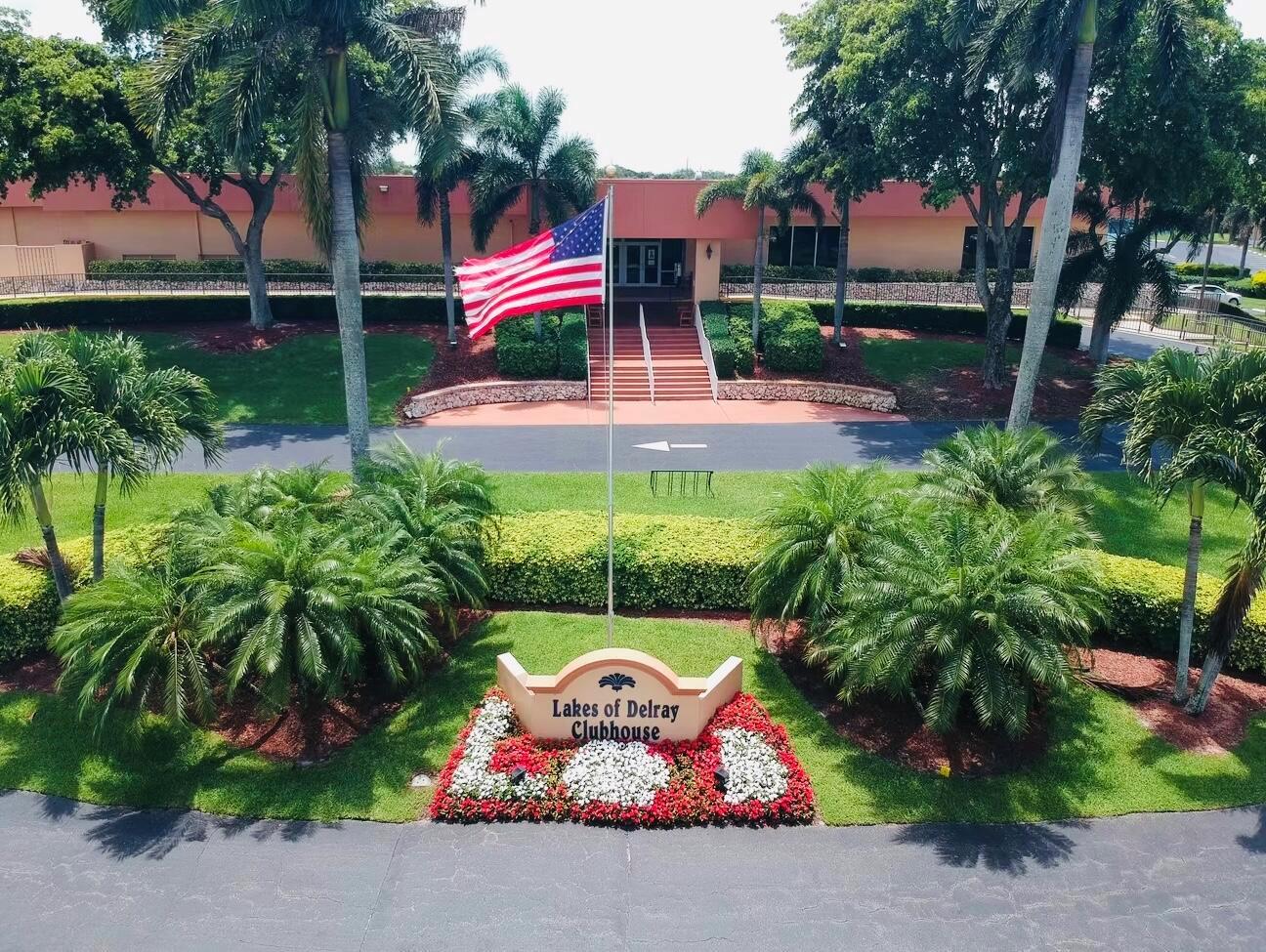 An aerial view of the Lakes of Delray clubhouse exterior with a flag and landscaping — 15072 Ashland Place S # 117, Delray Beach FL 33484, DELRAY BEACH, FL