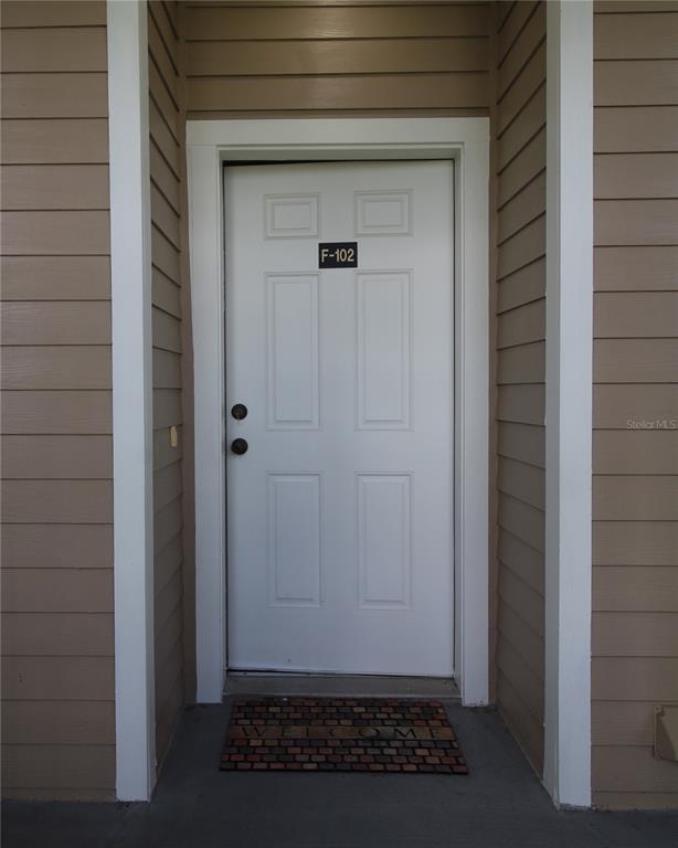 A white front door labeled F-102 is set within a tan, horizontal-sided exterior wall — 221 3RD AVENUE E UNIT F-102, HORSESHOE BEACH, FL