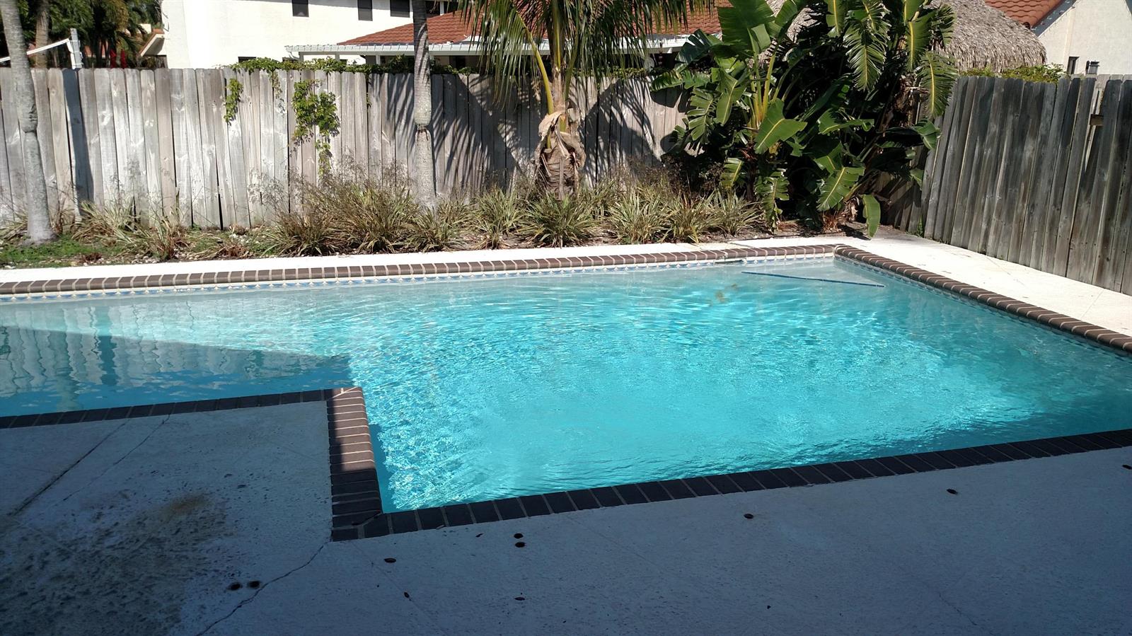A rectangular swimming pool with a brick border sits in a backyard with a wooden fence — 451 W Conference Drive, Boca Raton FL 33486, BOCA RATON, FL
